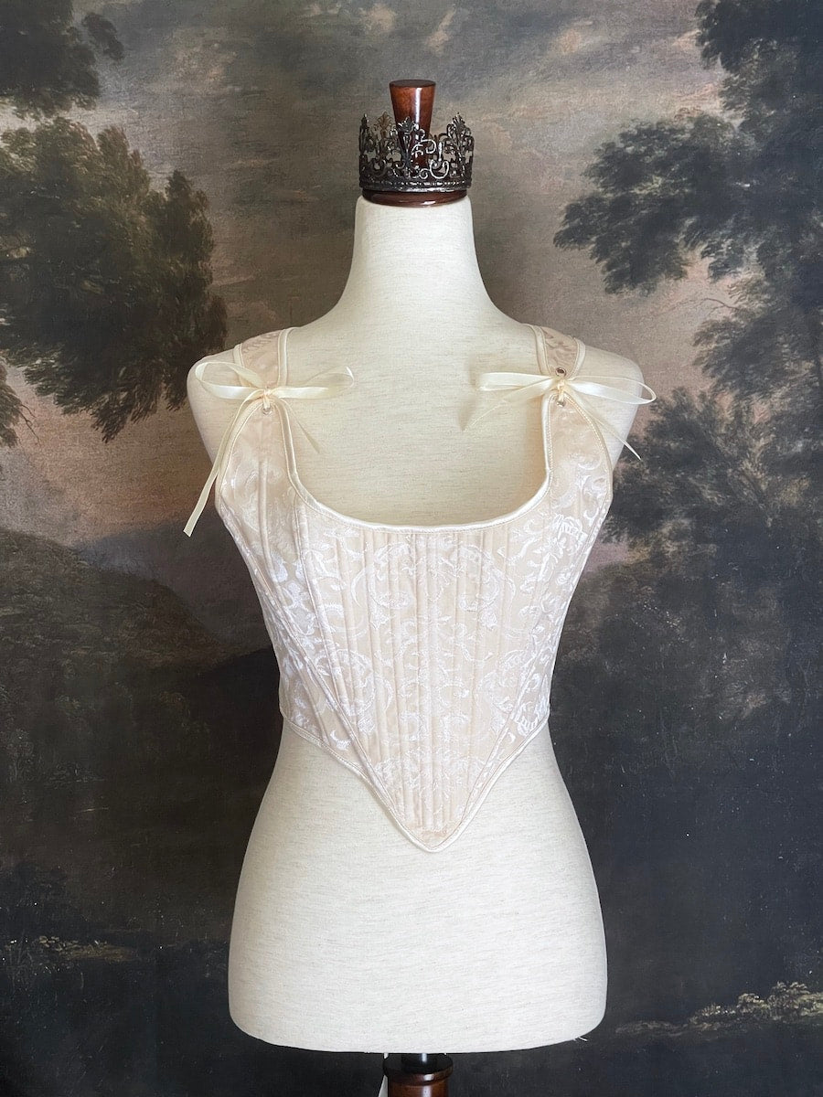 A Historically inspired Tudor, baroque, or renaissance era corset with champagne-ivory colored ornate jacquard fabric is pictured on a mannequin in front of a historic painted landscape backdrop.