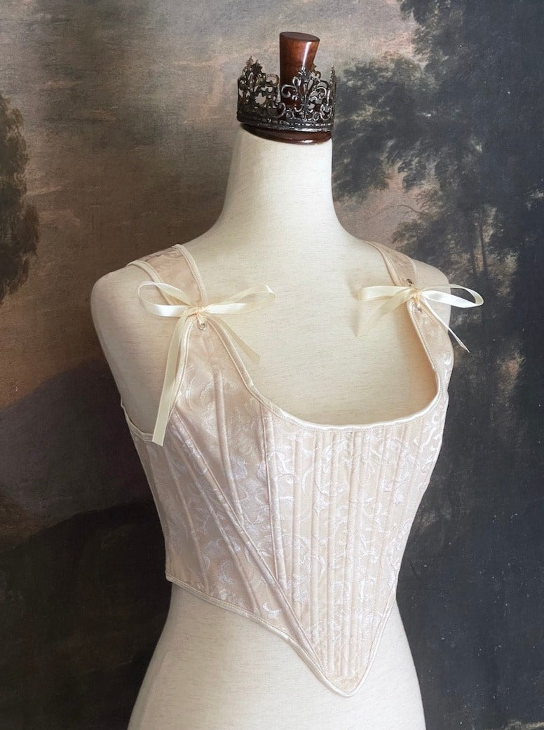A Historically inspired Tudor, baroque, or renaissance era corset with champagne-ivory colored ornate jacquard fabric is pictured on a mannequin in front of a historic painted landscape backdrop.