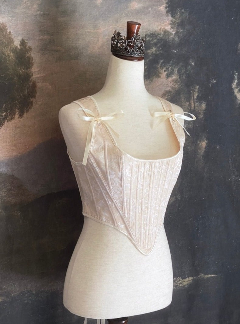 A Historically inspired Tudor, baroque, or renaissance era corset with champagne-ivory colored ornate jacquard fabric is pictured on a mannequin in front of a historic painted landscape backdrop.