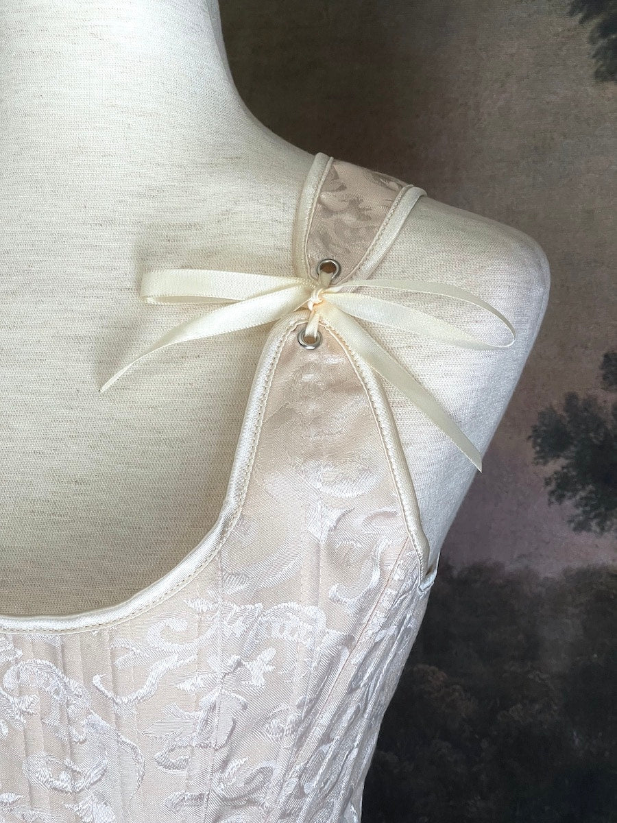 A Historically inspired Tudor, baroque, or renaissance era corset with champagne-ivory colored ornate jacquard fabric is pictured on a mannequin in front of a historic painted landscape backdrop.