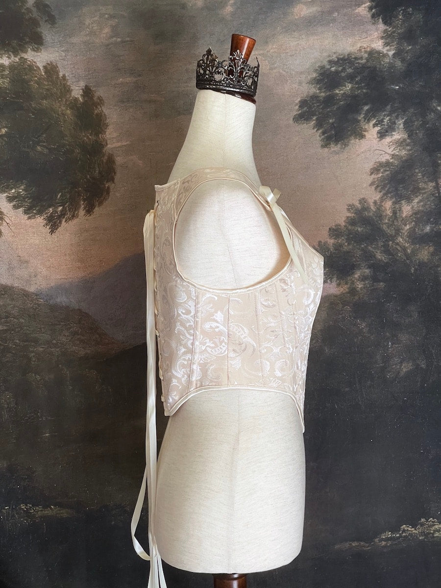 A Historically inspired Tudor, baroque, or renaissance era corset with champagne-ivory colored ornate jacquard fabric is pictured on a mannequin in front of a historic painted landscape backdrop.