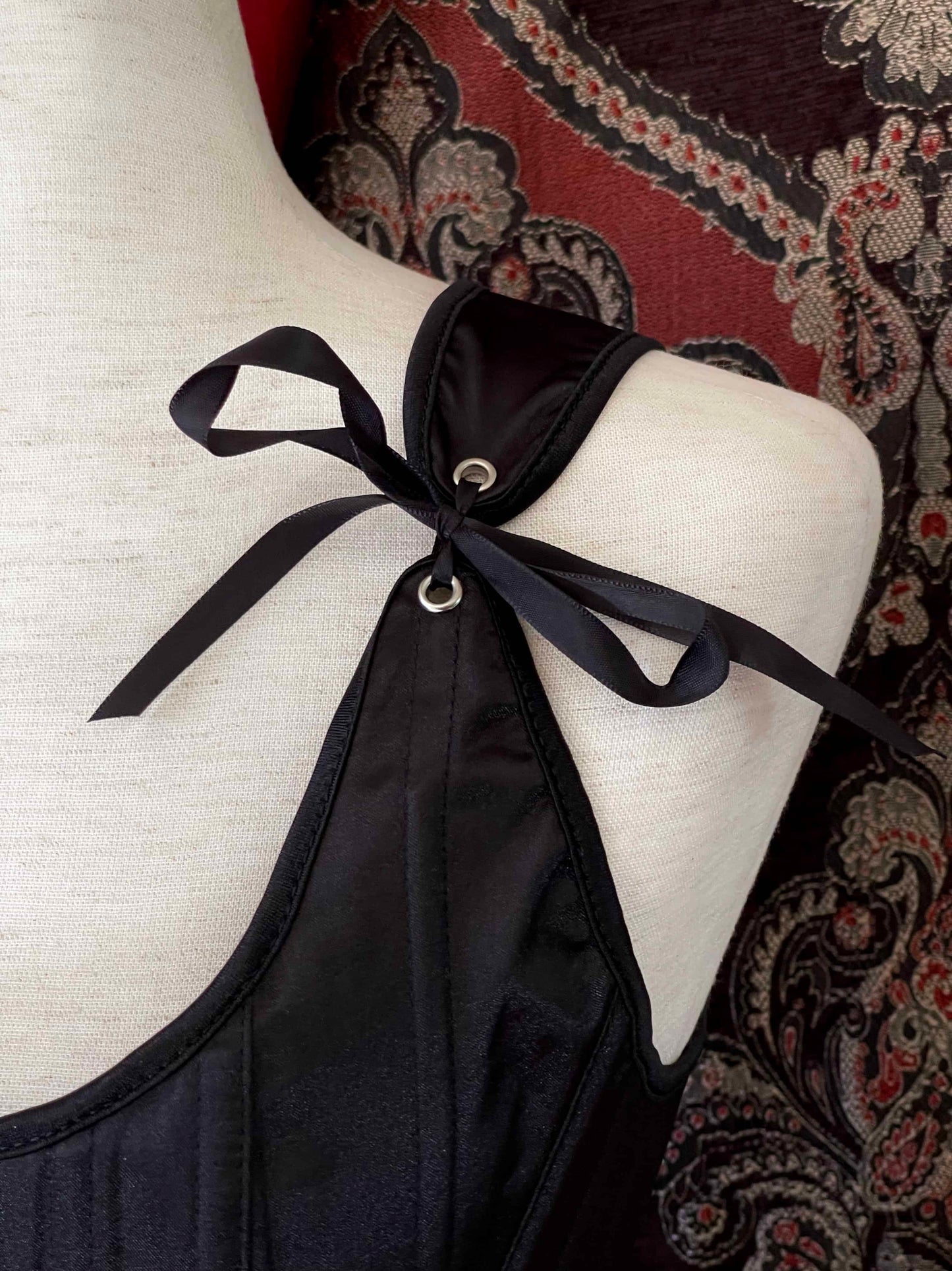 A Gothic Black Satin Renaissance Tudor Era Corset or Pair of stays is pictured on a mannequin in front of a historical Ornate Backdrop.