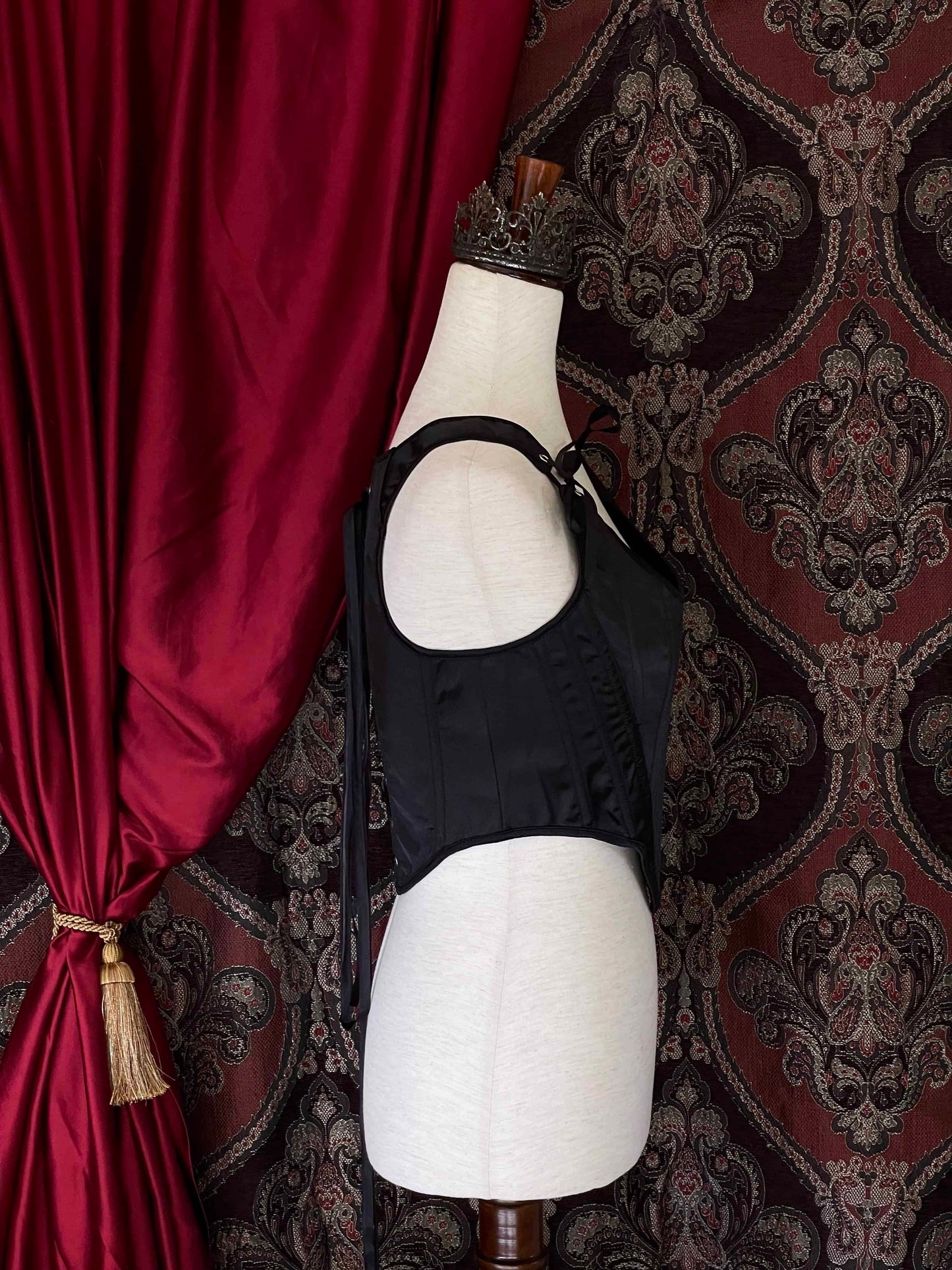 A Gothic Black Satin Renaissance Tudor Era Corset or Pair of stays is pictured on a mannequin in front of a historical Ornate Backdrop.