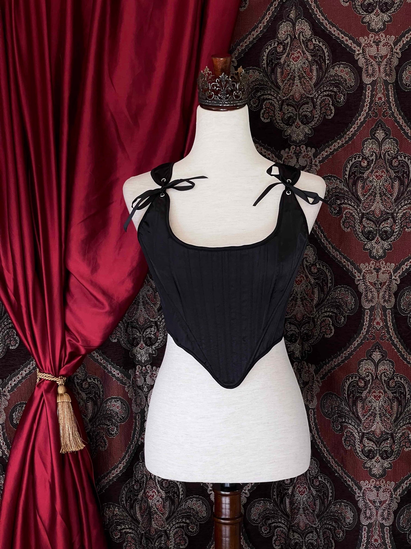 A Gothic Black Satin Renaissance Tudor Era Corset or Pair of stays is pictured on a mannequin in front of a historical Ornate Backdrop.