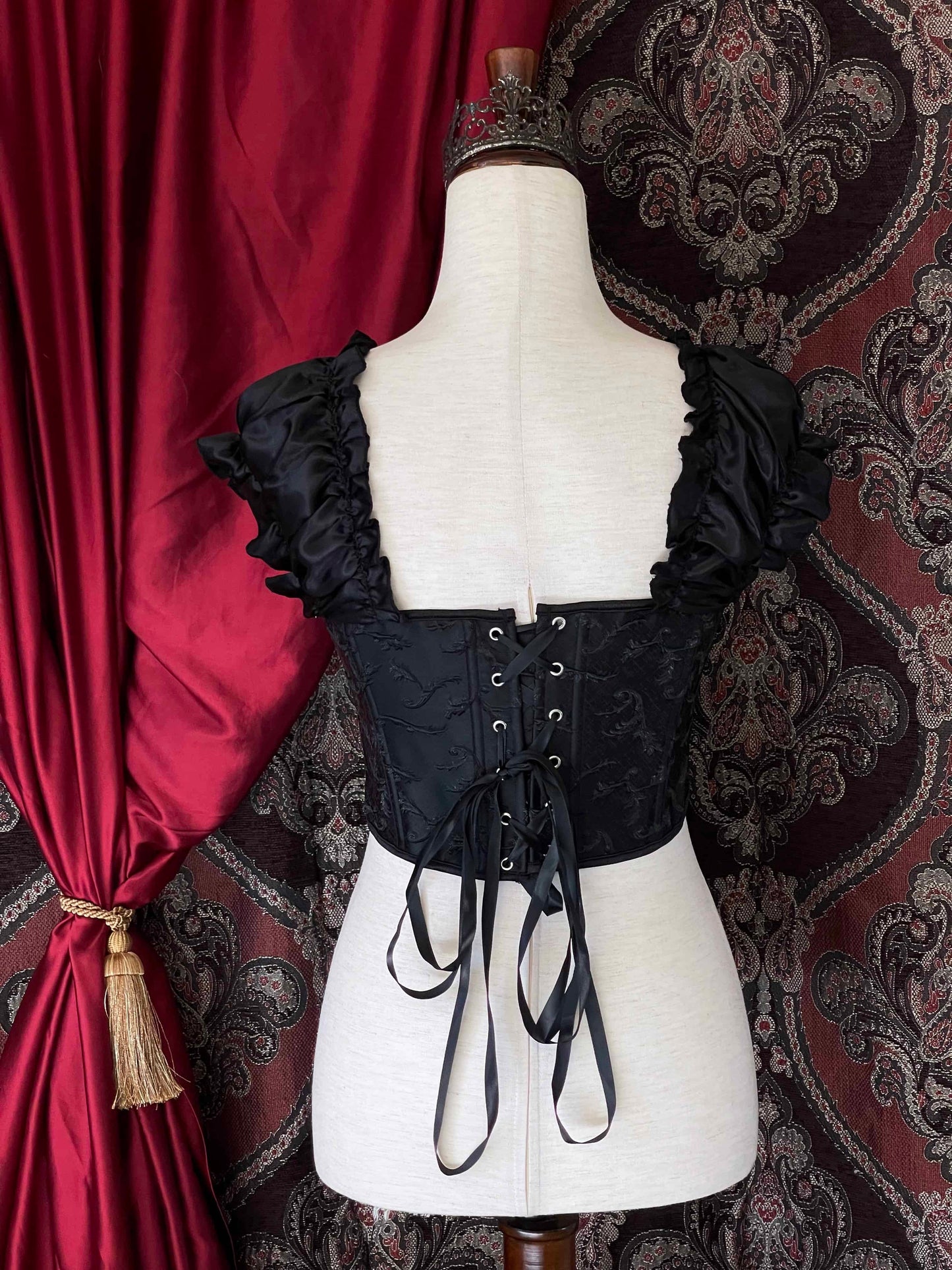 A historically inspired renaissance and baroque era corset made of black embroidered fabric and puff sleeves is pictured on a mannequin in front of an ornate backdrop.