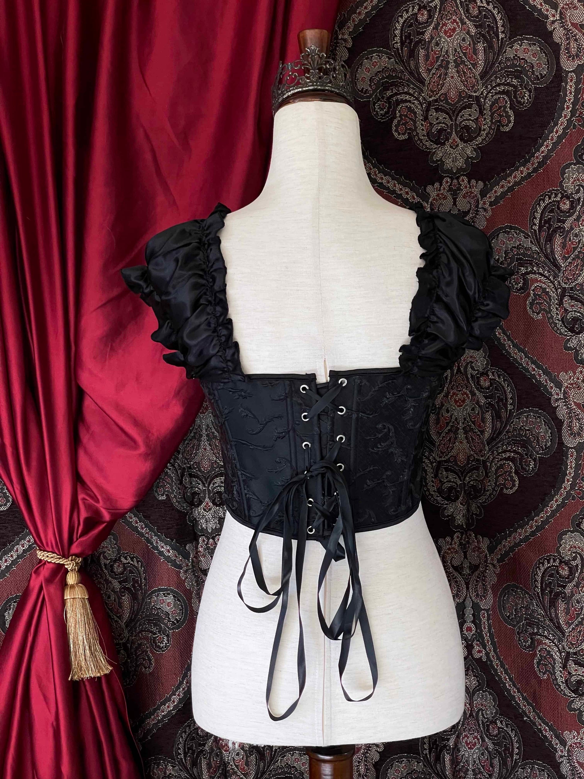 A historically inspired renaissance and baroque era corset  made of black embroidered fabric and puff sleeves is pictured on a mannequin in front of an ornate backdrop.