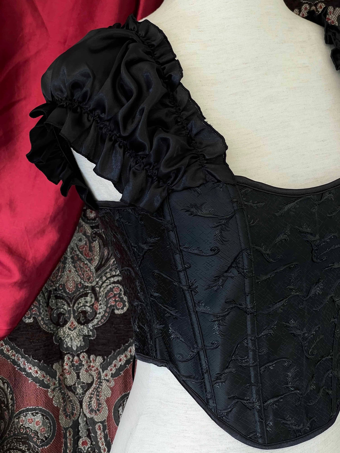 A historically inspired renaissance and baroque era corset made of black embroidered fabric and puff sleeves is pictured on a mannequin in front of an ornate backdrop.