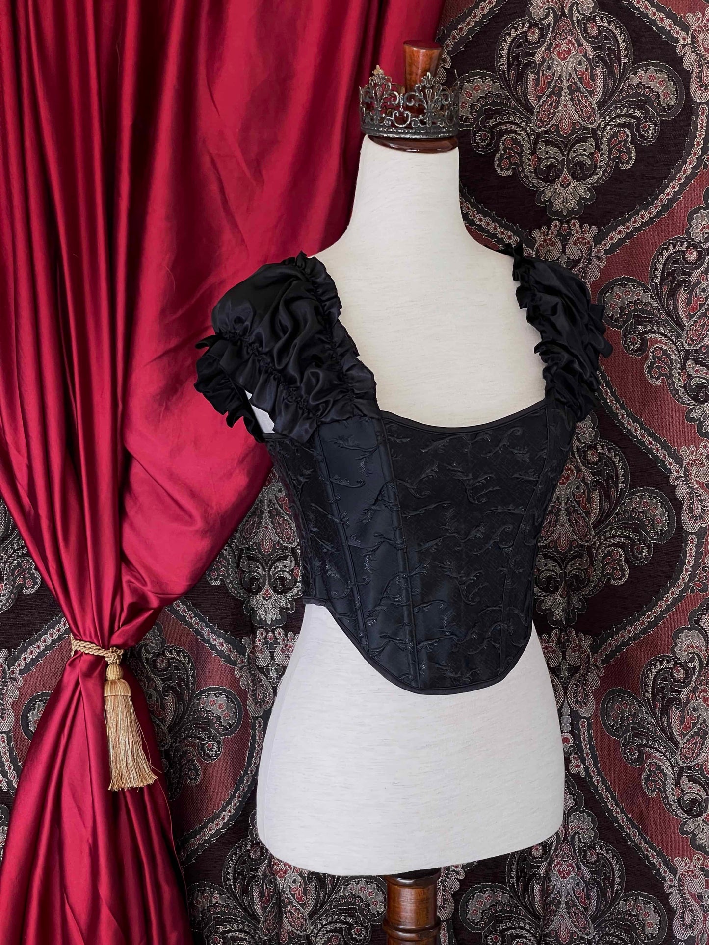 A historically inspired renaissance and baroque era corset made of black embroidered fabric and puff sleeves is pictured on a mannequin in front of an ornate backdrop.