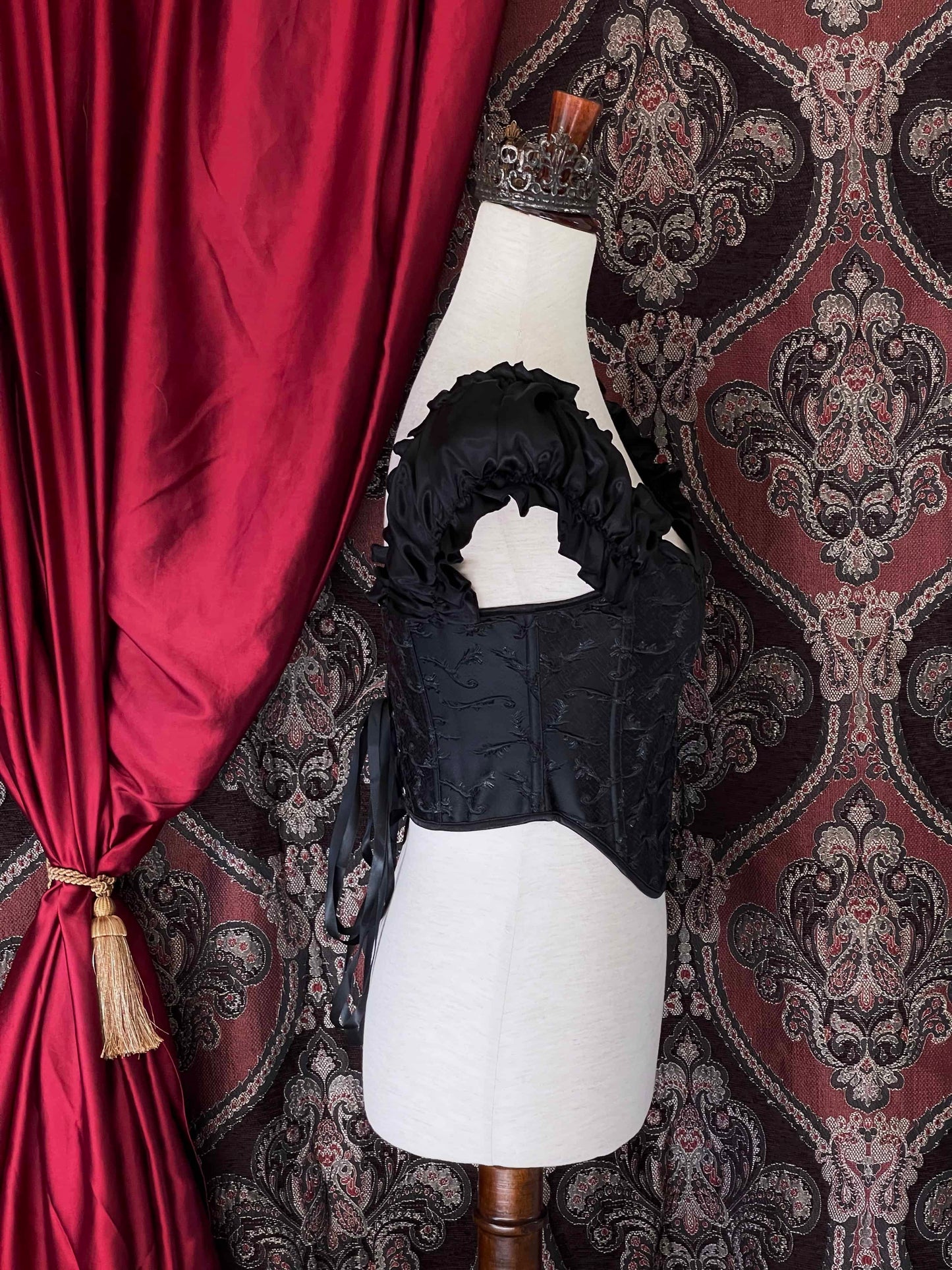 A historically inspired renaissance and baroque era corset made of black embroidered fabric and puff sleeves is pictured on a mannequin in front of an ornate backdrop.