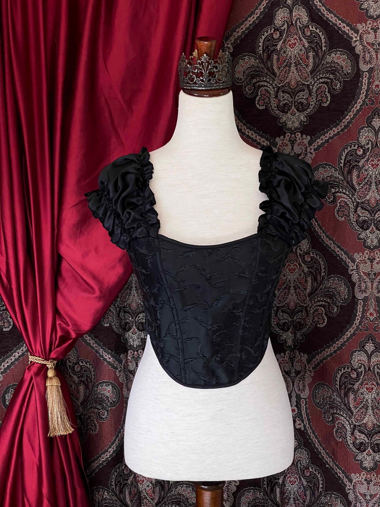 A historically inspired renaissance and baroque era corset made of black embroidered fabric and puff sleeves is pictured on a mannequin in front of an ornate backdrop.
