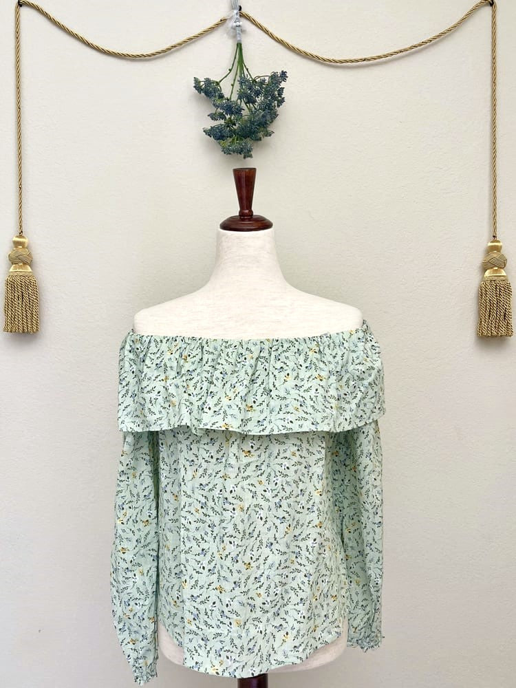A light mint green floral print off the shoulder blouse inspired by victorian era fashion is pictured on a mannequin in front of a floral bouquet and gold tassel.