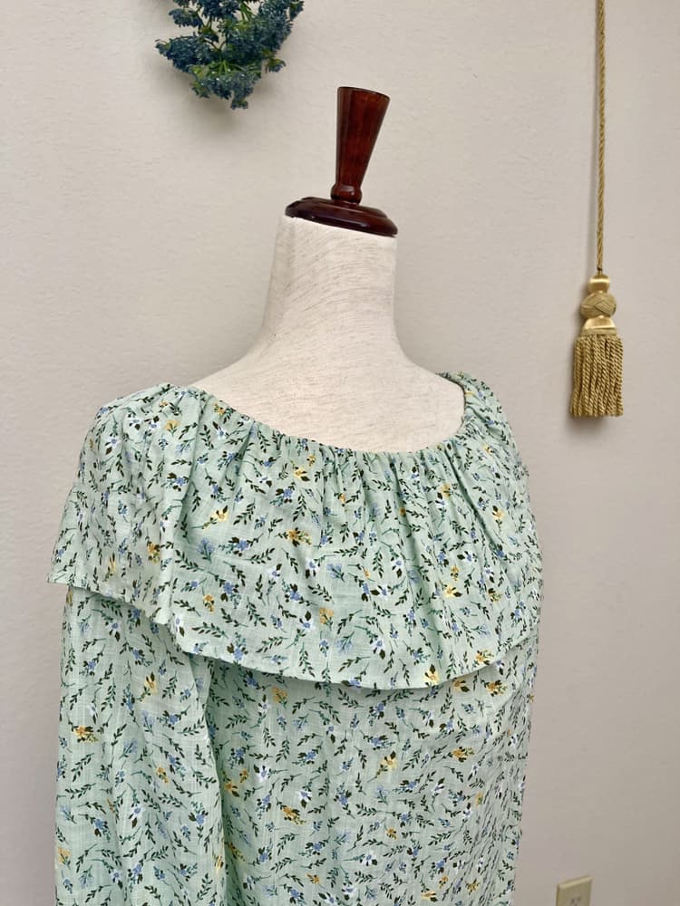 A light mint green floral print off the shoulder blouse inspired by victorian era fashion is pictured on a mannequin in front of a floral bouquet and gold tassel.