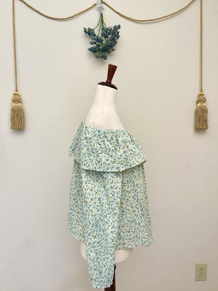 A light mint green floral print off the shoulder blouse inspired by victorian era fashion is pictured on a mannequin in front of a floral bouquet and gold tassel.