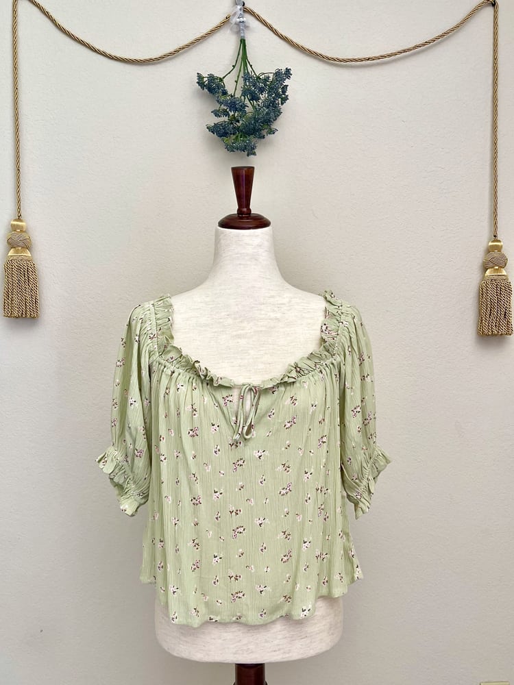 A Sage Green Historically Inspired Short Sleeve Peasant Blouse with Tie Front Ruched Neckline is pictured on a mannequin in front of a classical background.