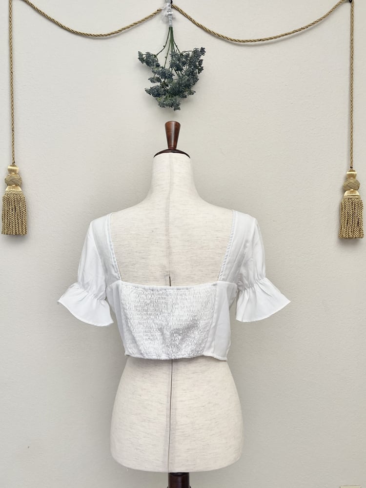 A white edwardian era inspired cropped blouse with ruffle sleeves is pictured on a mannequin in front of a classical background.