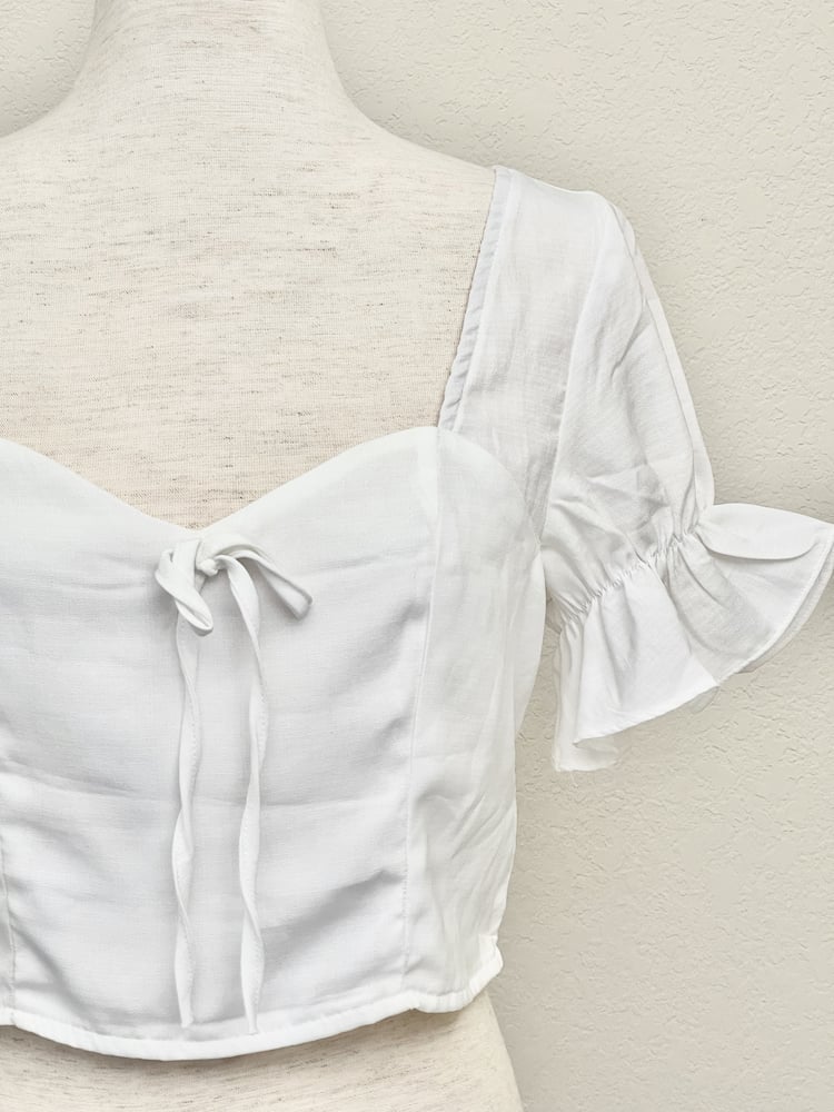 A white edwardian era inspired cropped blouse with ruffle sleeves is pictured on a mannequin in front of a classical background.