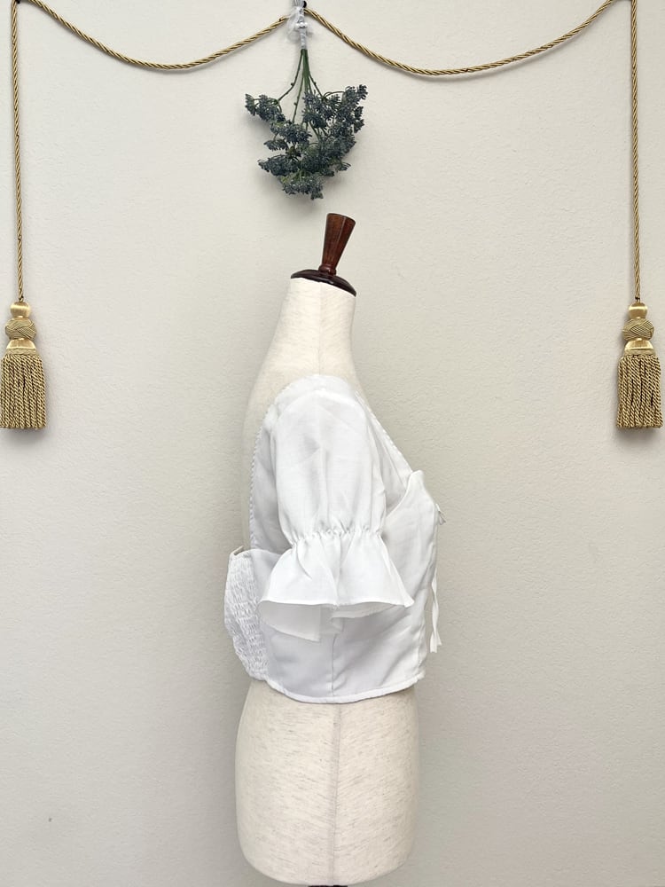 A white edwardian era inspired cropped blouse with ruffle sleeves is pictured on a mannequin in front of a classical background.