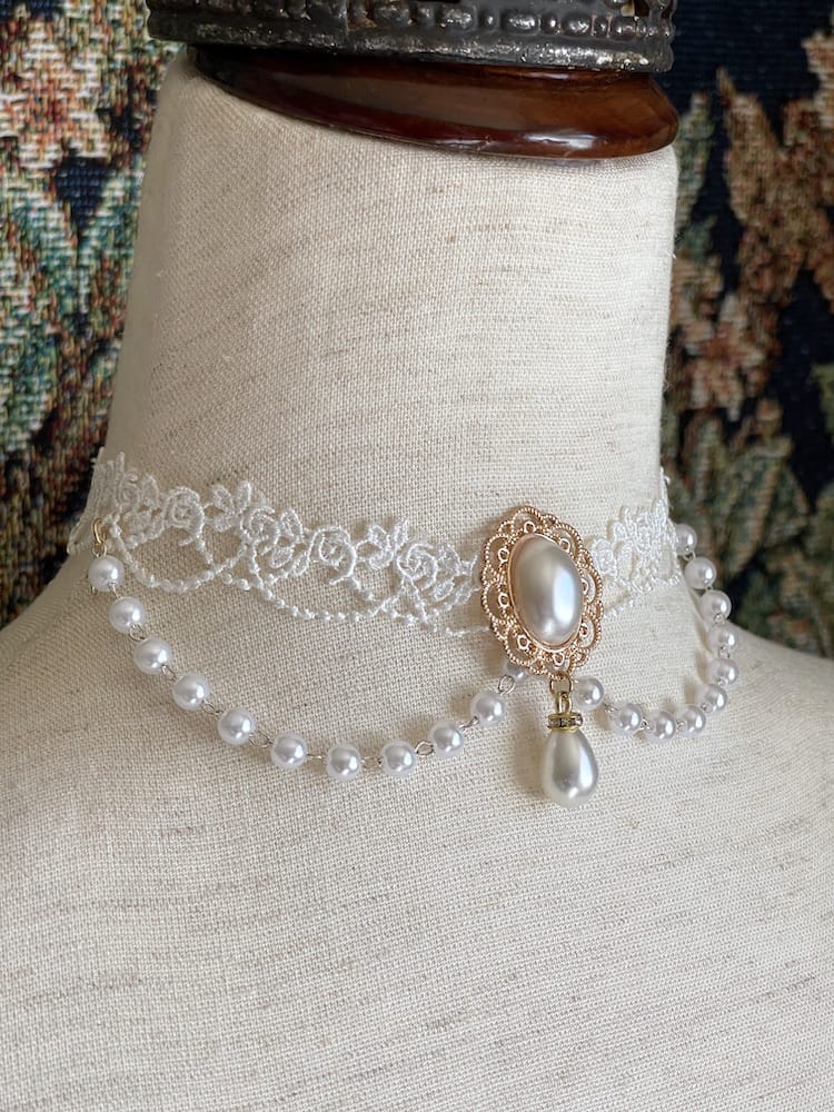A Rococo or Regency era historically inspired pearl and floral lace choker necklace is pictured on a mannequin in front of a historic tapestry.