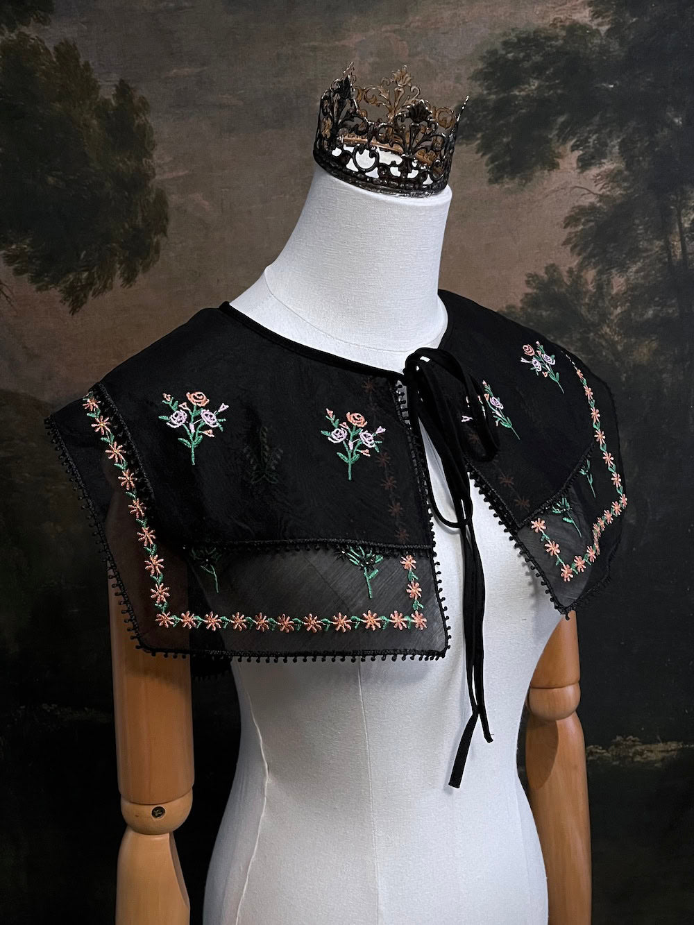 Mannequin wearing a black embroidered cape collar with floral designs against a dark art history painting background
