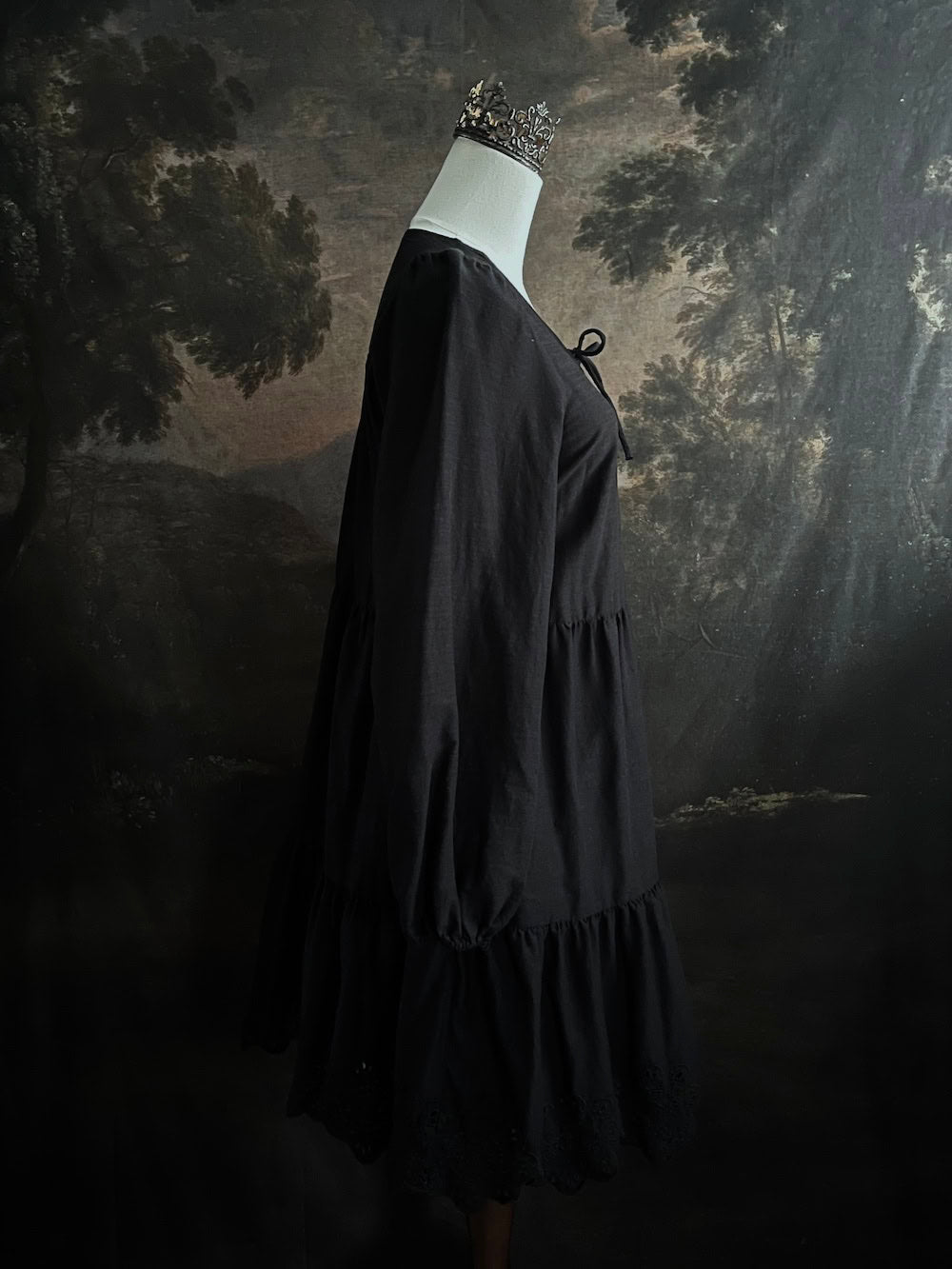Historically inspired witchy gothic black tiered mini dress with corset lace up bodice and long bishop sleeves on a mannequin in front of a dark art history backdrop