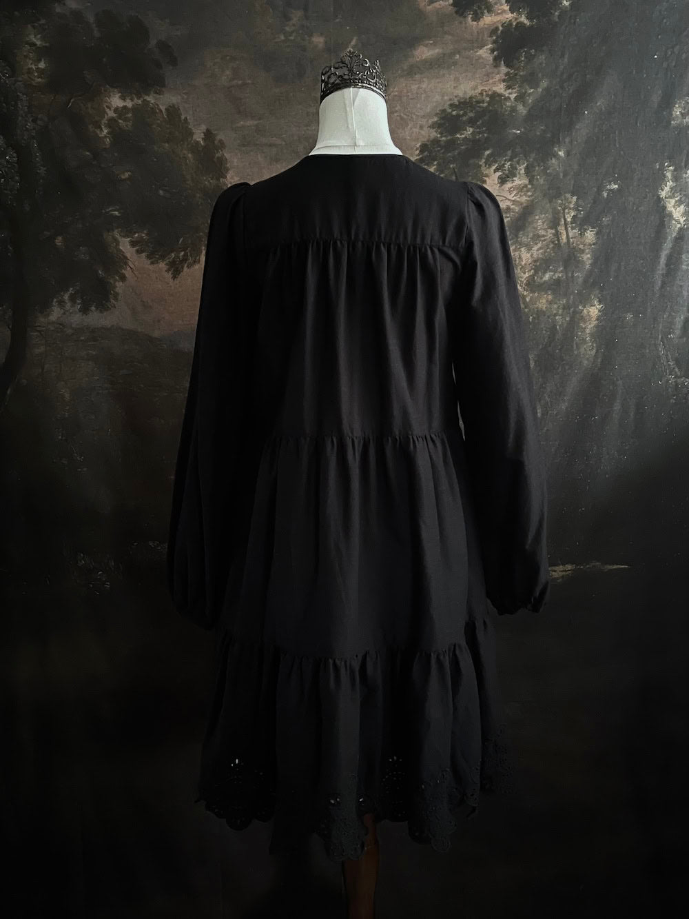Historically inspired witchy gothic black tiered mini dress with corset lace up bodice and long bishop sleeves on a mannequin in front of a dark art history backdrop