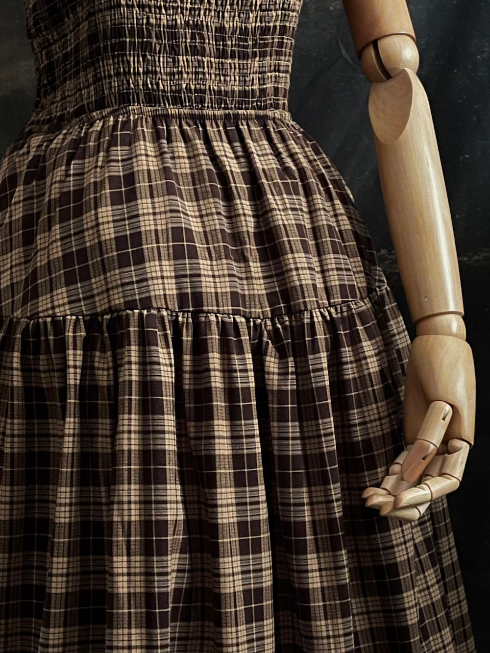 up close of a Mannequin wearing a historical fashion inspired brown tartan plaid maxi dress against a dark art history background