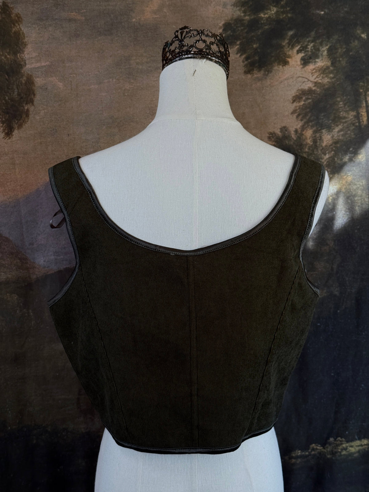 A historically inspired dark olive moss green corduroy corset top with brown lace-up front ties and shoulder ties, displayed on a mannequin against a scenic landscape painting backdrop.