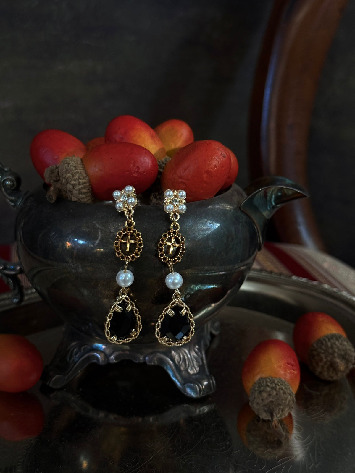 A pair of renaissance inspired ornate gold and black drop earrings with white pearl-like beads, hanging from a dark antique pot filled with red acorns on a reflective silver tray.