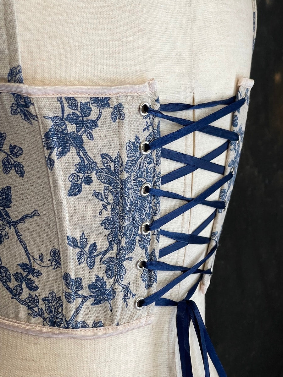 A rococo and regency era inspired blue and ivory colored floral toile de jouy corset pair of stays in front of a historical patterned wallpaper.