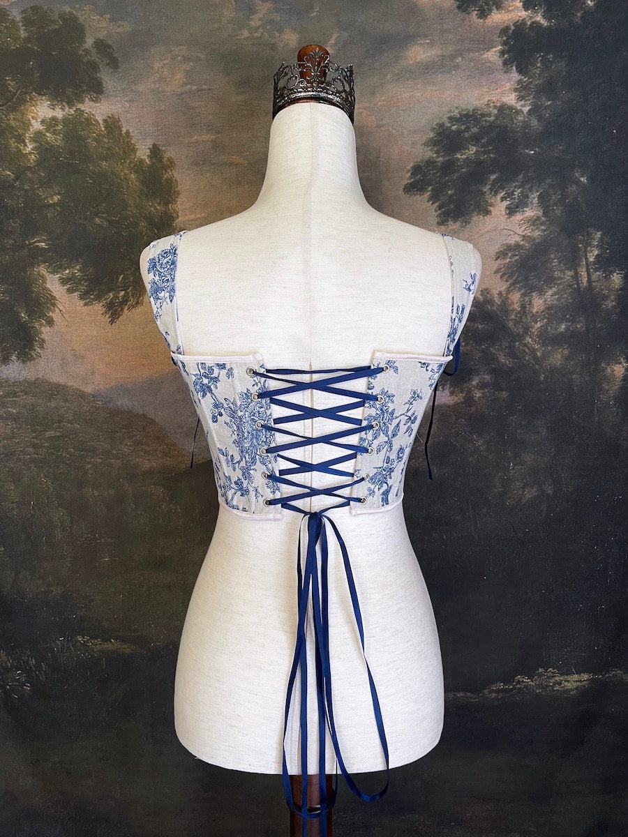A rococo and regency era inspired blue and ivory colored floral toile de jouy corset pair of stays in front of a historical patterned wallpaper.