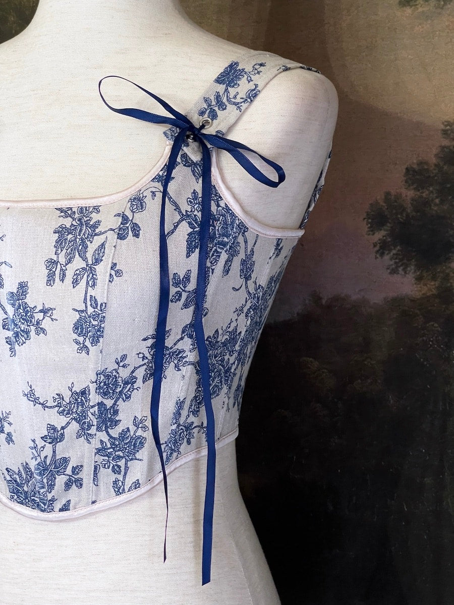 A rococo and regency era inspired blue and ivory colored floral toile de jouy corset pair of stays in front of a historical patterned wallpaper.