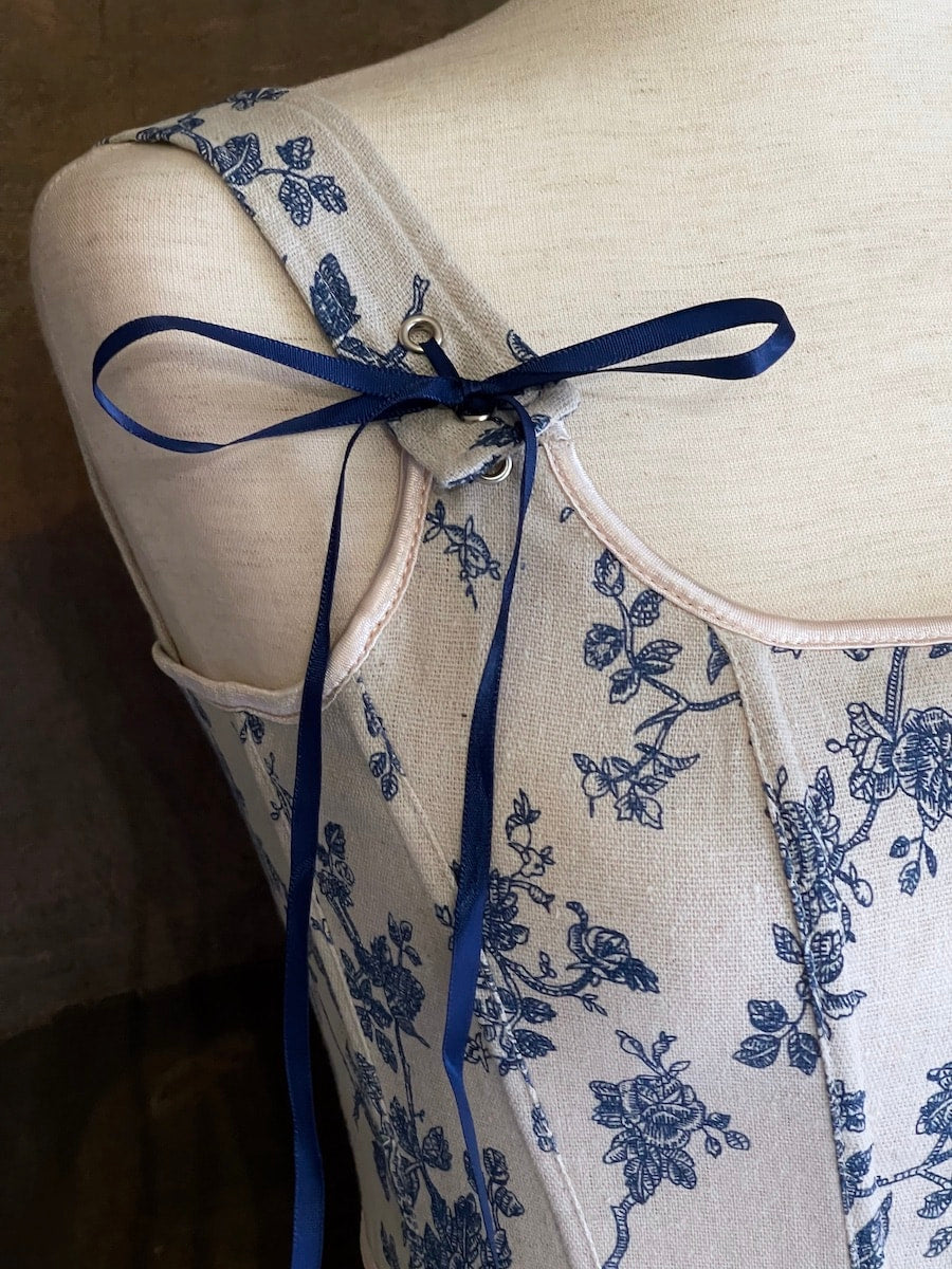 A rococo and regency era inspired blue and ivory colored floral toile de jouy corset pair of stays in front of a historical patterned wallpaper.