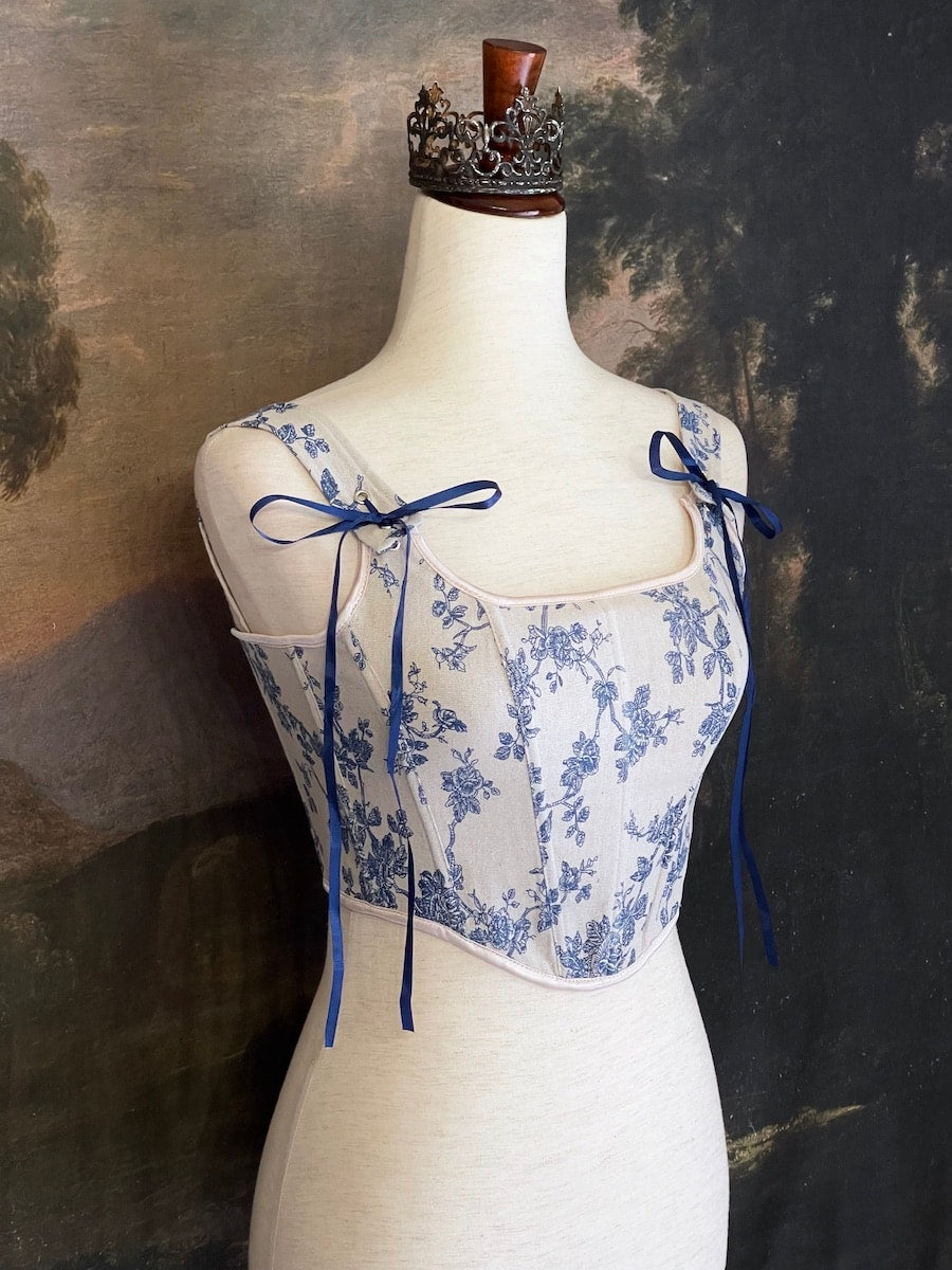 A rococo and regency era inspired blue and ivory colored floral toile de jouy corset pair of stays in front of a historical patterned wallpaper.