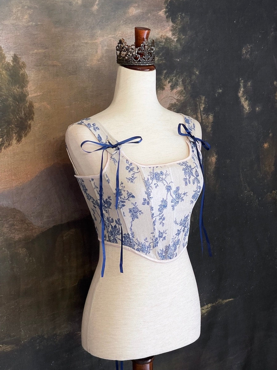 A rococo and regency era inspired blue and ivory colored floral toile de jouy corset pair of stays in front of a historical patterned wallpaper.
