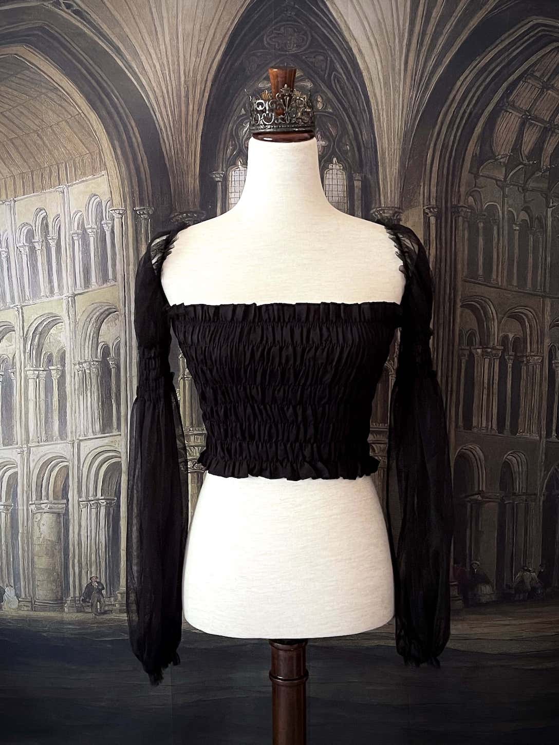 A historically inspired gothic black smocked blouse with mesh juliet sleeves is pictured on a mannequin in front of a medieval gothic cathedral backdrop.