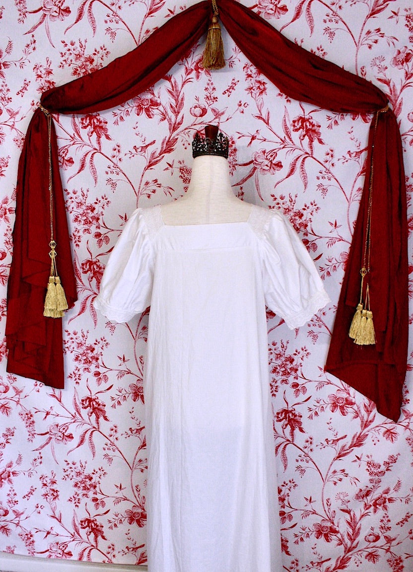 A historically inspired regency style short sleeve chemise dress with pin-tuck pleats and lace trim in front of a toile backdrop.