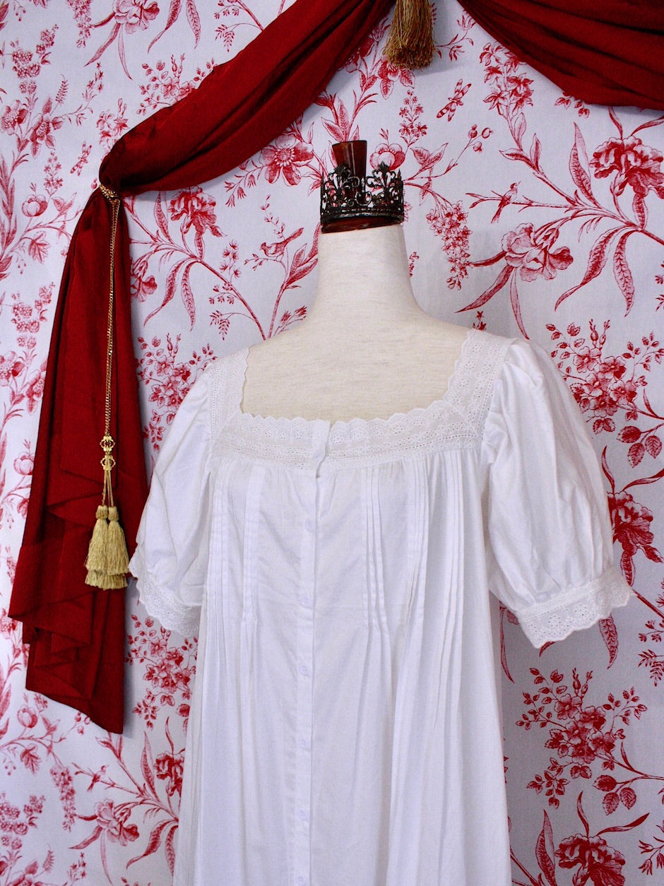 A historically inspired regency style short sleeve chemise dress with pin-tuck pleats and lace trim in front of a toile backdrop.