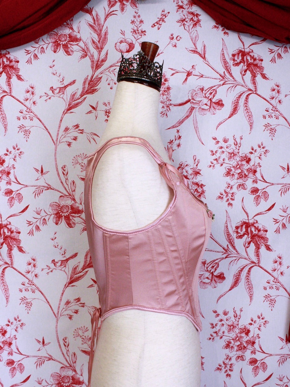 A pair of Balletcore Renaissance, Baroque, Rococo, Regency, Victorian Era Corset Stays in Ballet Pastel Pink with Rosette Appliques in front of a toile backdrop.