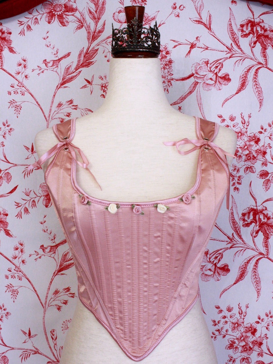 A pair of Balletcore Renaissance, Baroque, Rococo, Regency, Victorian Era Corset Stays in Ballet Pastel Pink with Rosette Appliques in front of a toile backdrop.