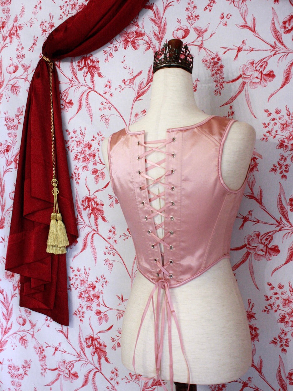 A pair of Balletcore Renaissance, Baroque, Rococo, Regency, Victorian Era Corset Stays in Ballet Pastel Pink with Rosette Appliques in front of a toile backdrop.