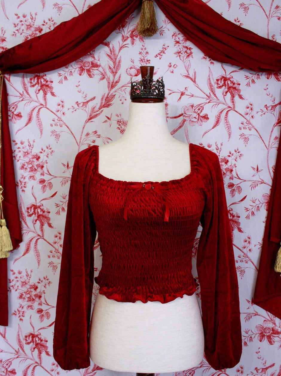 A Historically Inspired Renaissance to Victorian Era Red Velvet Smocked Blouse with Rosette Bow Applique in front of a toile backdrop.