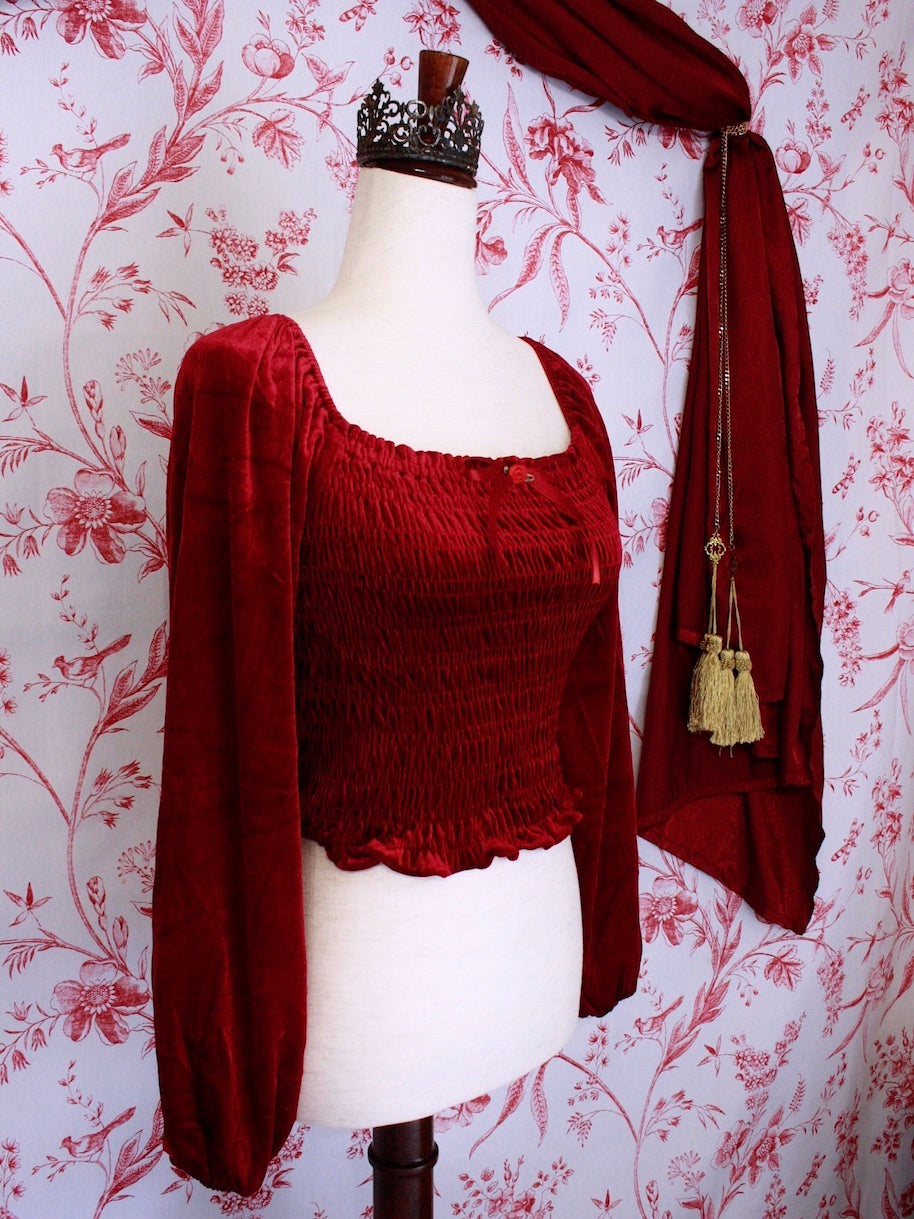 A Historically Inspired Renaissance to Victorian Era Red Velvet Smocked Blouse with Rosette Bow Applique in front of a toile backdrop.