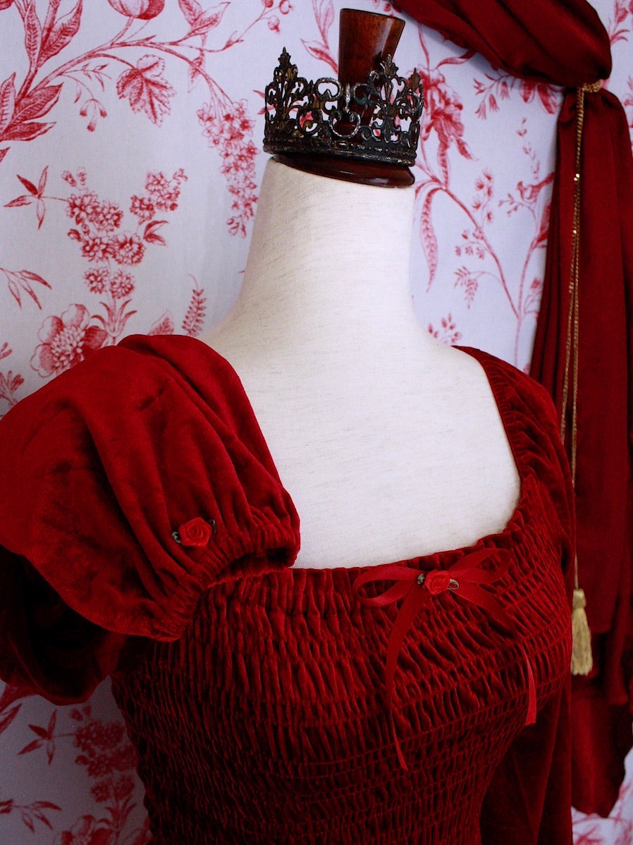 A Historically Inspired Renaissance to Victorian Era Red Velvet Smocked Blouse with Rosette Bow Applique in front of a toile backdrop.