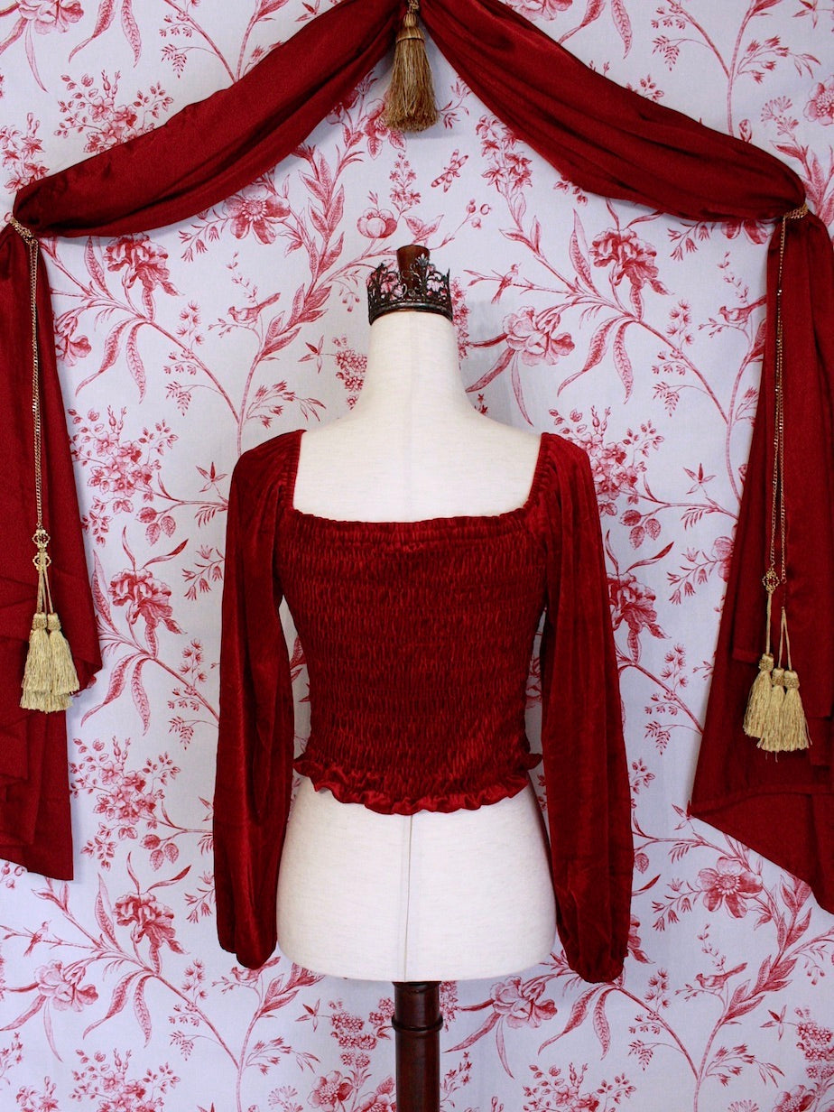 A Historically Inspired Renaissance to Victorian Era Red Velvet Smocked Blouse with Rosette Bow Applique in front of a toile backdrop.