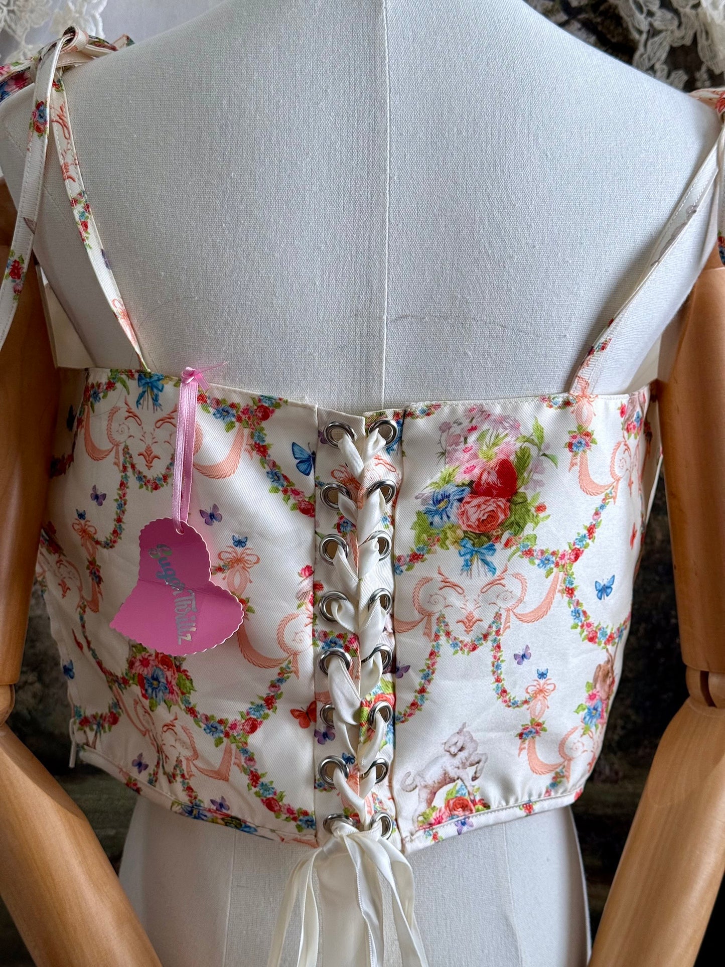 A historically-inspired 1700s rococo era corset top in an ivory floral printed satin is pictured on a mannequin in front of an art history backdrop.