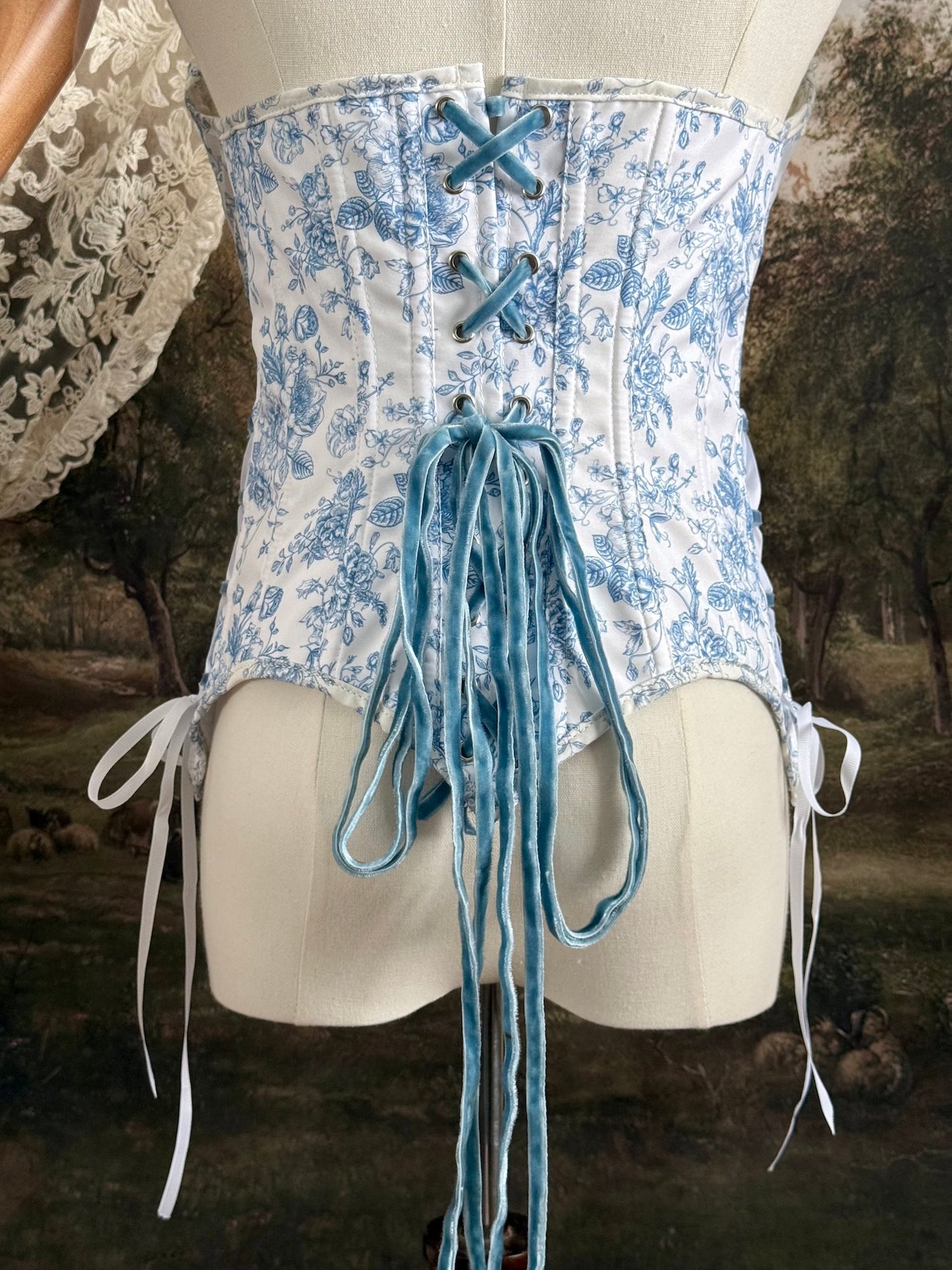 A historically-inspired vintage style overbust bustier corset in white and blue floral toile print, with a pointed basque waist, velvet lacing ties, lace trim, and a ruched bust detail, pictured on a mannequin. Back details.