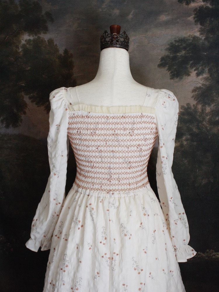 A Historically inspired rococo and regency Ivory bishop sleeve smocked tea gown with peach contrast stitching is pictured on a mannequin in front of a historical painting backdrop.