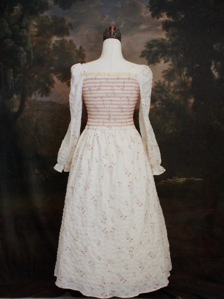 A Historically inspired rococo and regency Ivory bishop sleeve smocked tea gown with peach contrast stitching is pictured on a mannequin in front of a historical painting backdrop.