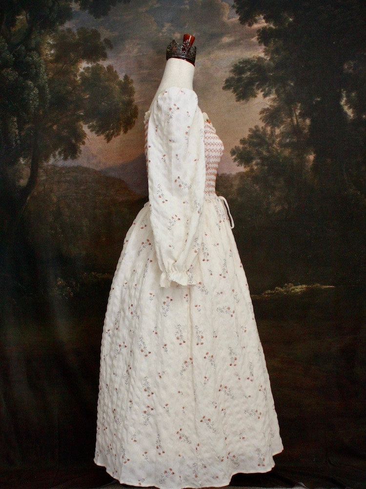 A Historically inspired rococo and regency Ivory bishop sleeve smocked tea gown with peach contrast stitching is pictured on a mannequin in front of a historical painting backdrop.