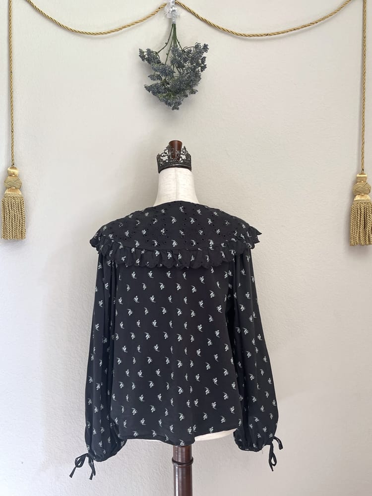 A black floral print historically inspired baroque blouse with large lace trimmed collar and long bishop sleeves is pictured on a mannequin from the back.